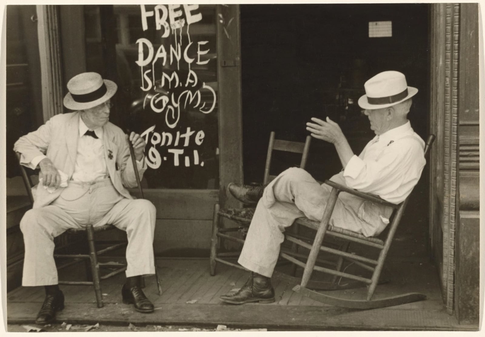 Two men sitting opposite each other on a porch, one gesturing mid-explanation