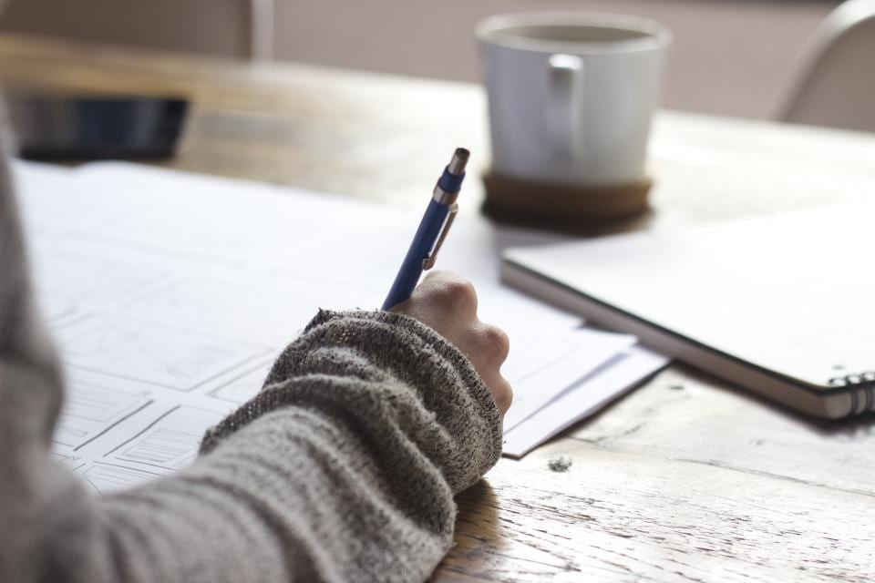 A hand writing on loose paper at a wooden desk beside a mug, in soft daylight